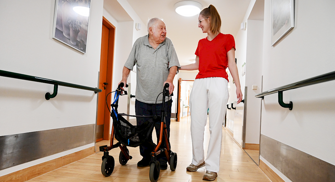 Eine AWO-Mitarbeiterin begleitet einen Klienten mit Rollator durch den Gang.