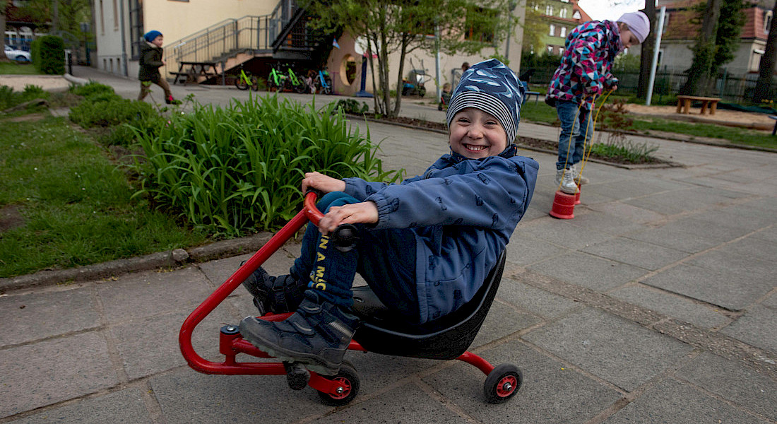 Ein Kind fährt lachend mit einem roten Dreirad über den Spielplatz der Kita.