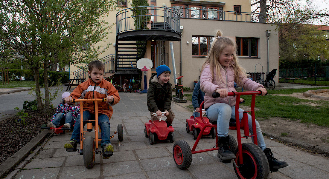 Mehrere Kinder fahren fröhlich mit Dreirädern und Fahrzeugen auf dem Hof der Kita.