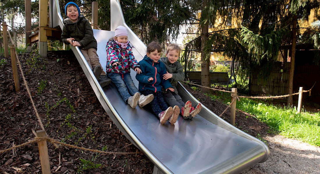 Kinder rutschen fröhlich auf einer langen Metallrutsche im Kita-Garten.