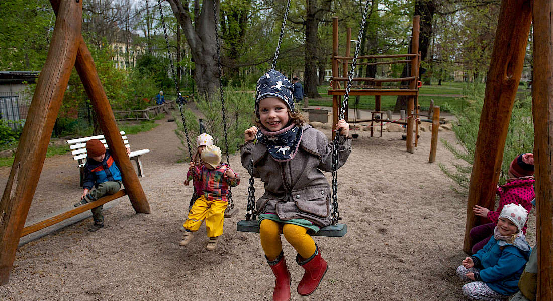 Fröhliche Kinder schaukeln und spielen gemeinsam auf dem Spielplatz der Kita. Im Mittelpunkt sitzt ein lächelndes Kind mit Mütze auf einer Schaukel.