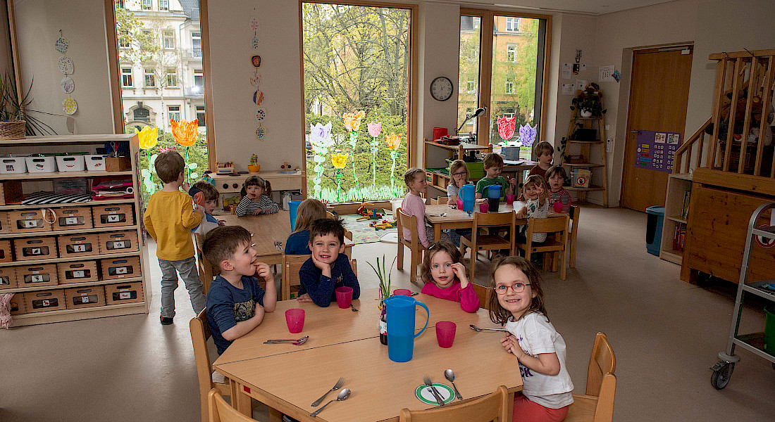 Fröhliche Kinder sitzen zusammen an Tischen in einem Gruppenraum der Kita und warten auf ihr Essen.