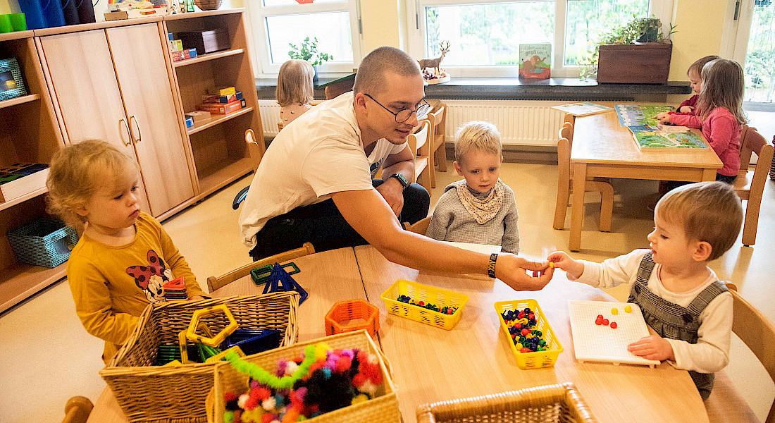 Ein Erzieher spielt mit Kindern an einem Tisch mit Holzspielzeug und Bastelmaterialien.