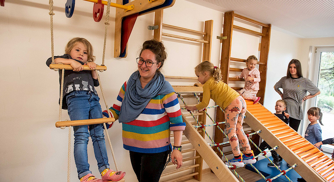 Ein Kind balanciert mit Unterstützung über eine Kletterbrücke im Turnraum. Andere Kinder hangeln sich an Netzen und Strickleitern entlang.