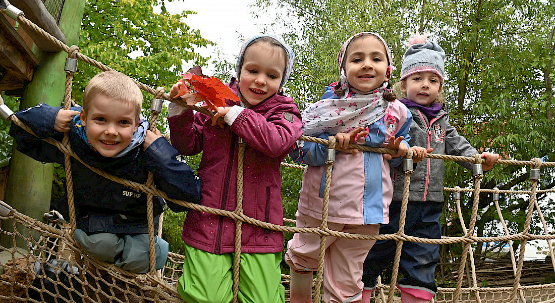 Vier Kinder stehen auf einer Hängebrücke im Außengelände und schauen durch und über das Netz der Brücke.
