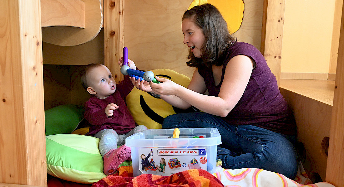 Ein Kleinkind und eine Fachkraft sitzen auf einer Decke und spielen gemeinsam mit Spielmaterial.