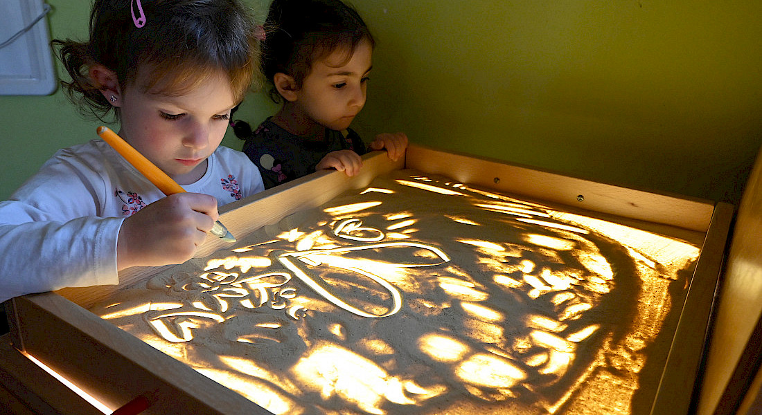 Zwei Kinder spielen konzentriert mit Licht und Sand auf einem Leuchttisch.