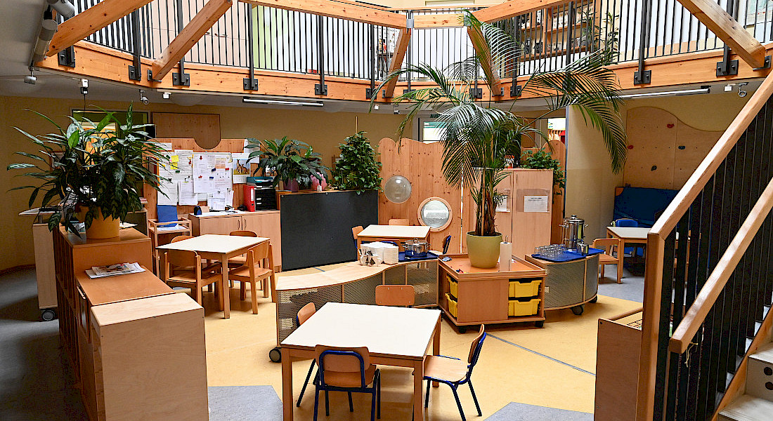 Blick in einen hellen Raum im Zentrum der Kita mit Tischen, Pflanzen und Galerieebene.
