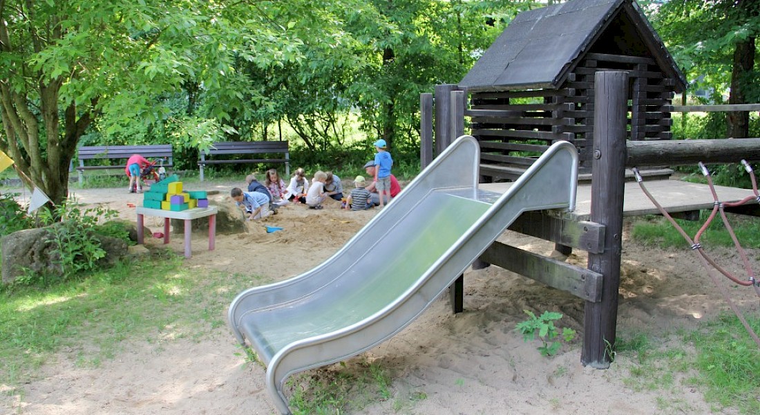 Ein Spielplatz mit Rutsche und schattigem Sitzbereich unter Bäumen.
