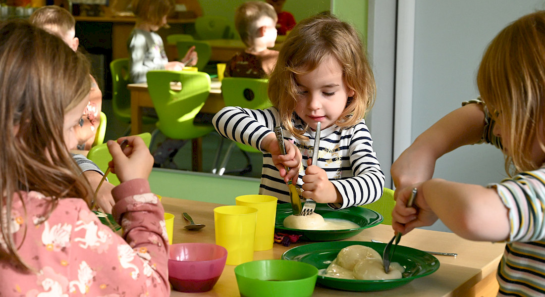 Kinder essen gemeinsam am Tisch in einem hellen Gruppenraum.