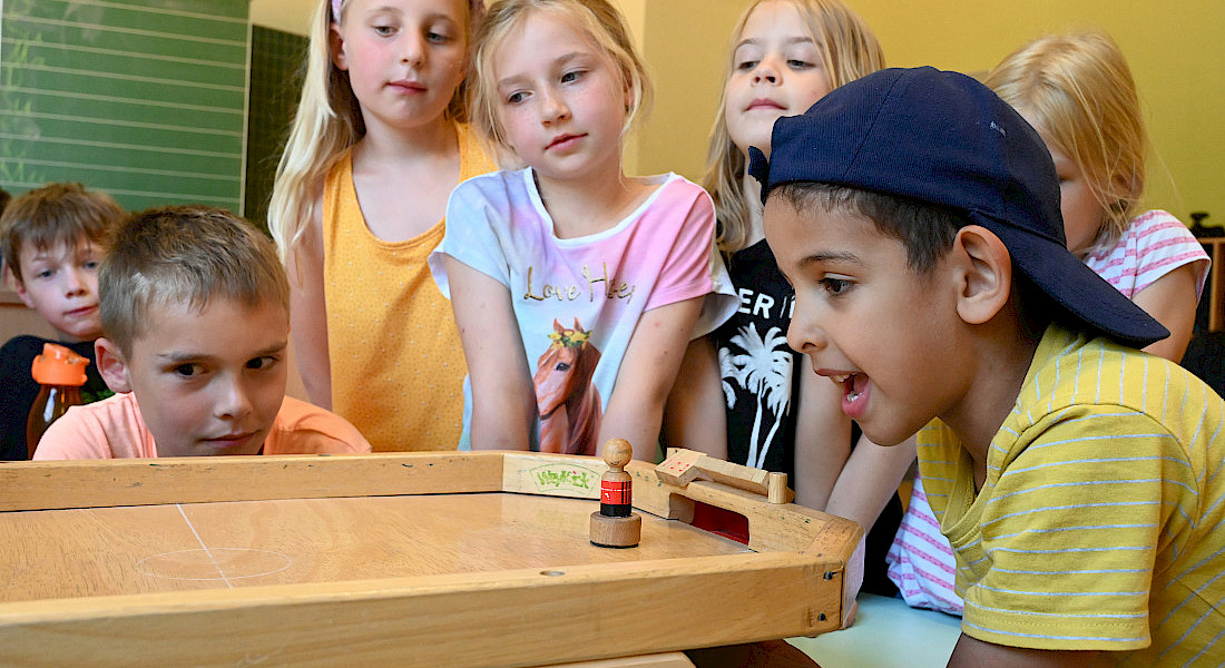 Mehrere Kinder beobachten gespannt ein Spiel auf einem Holzbrett mit Tor.
