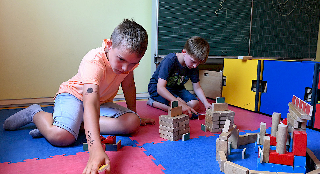 Zwei Kinder bauen mit Holzklötzen auf einer gepolsterten Spielfläche.