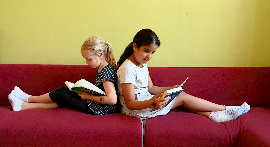 Zwei Kinder lesen nebeneinander auf einem Sofa in einem hellen Raum.