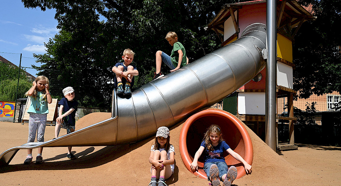 Kinder sitzen und klettern auf einem Spielgerät mit Röhrenrutsche im Außengelände.