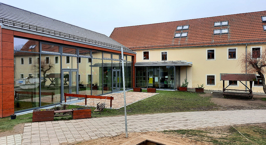 Ansicht eines hellen Kita-Gebäudes mit überdachtem Eingangsbereich und Garten.