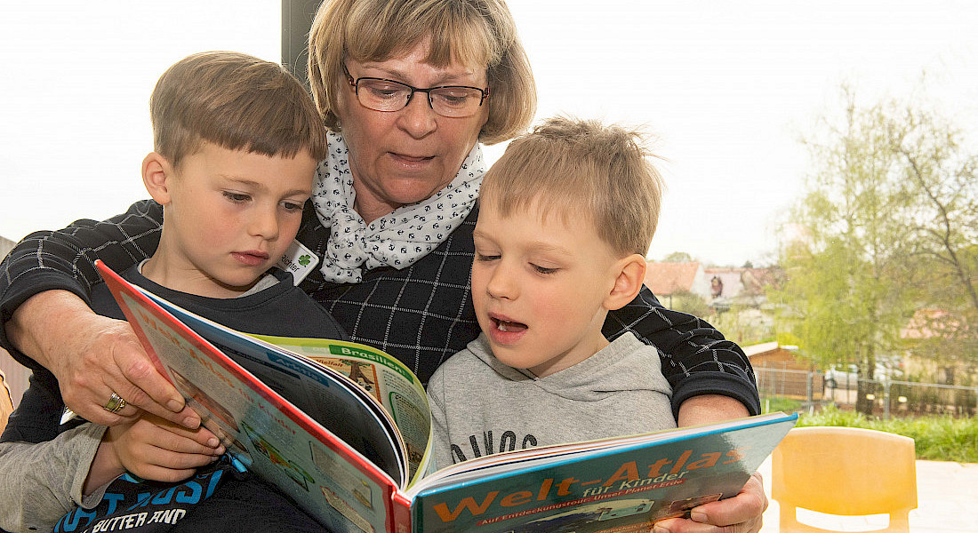 Zwei Kinder schauen mit einer Fachkraft gemeinsam in ein Bilderbuch.
