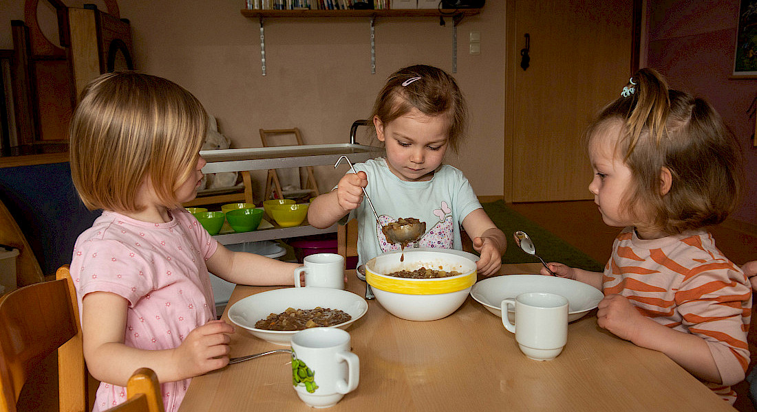 Kinder sitzen an einem Tisch und schöpfen eigenständig Essen in Schüsseln.