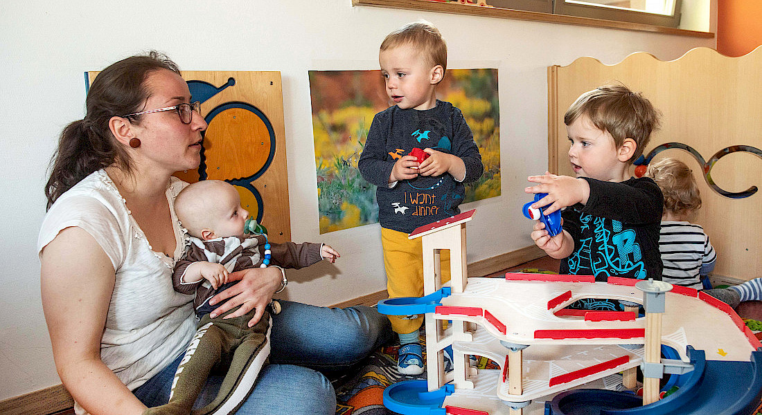 Kleinkinder spielen zusammen im Gruppenraum mit Parkhaus, eine erwachsene Person sitzt dabei.