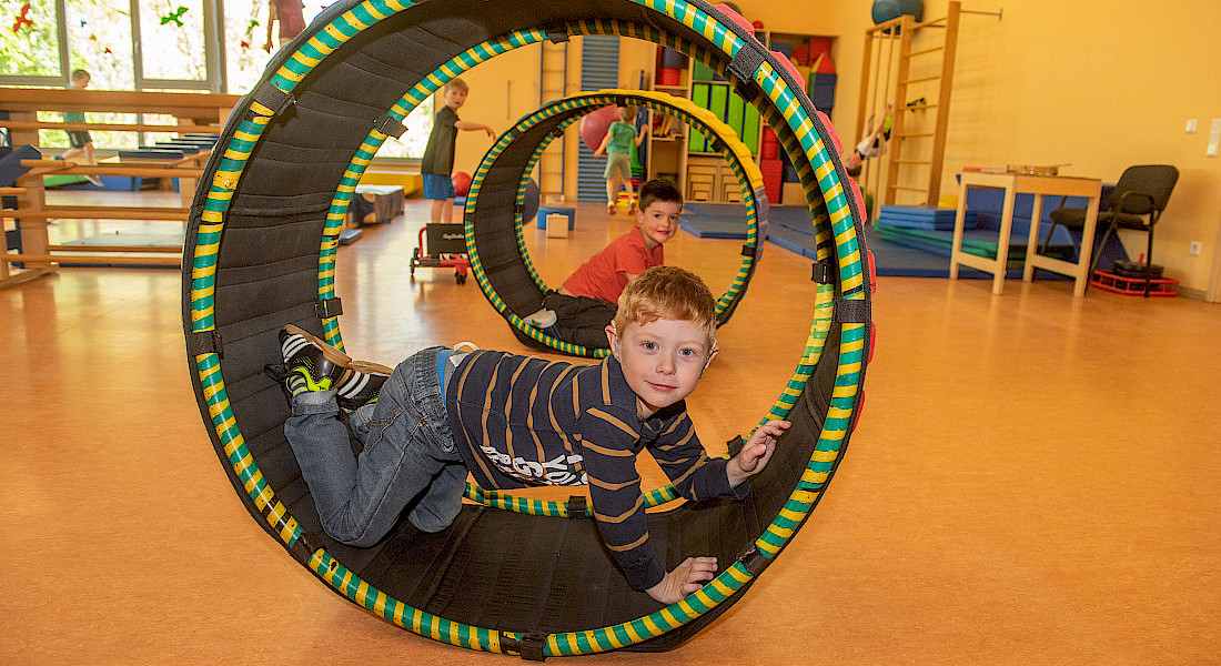 Kinder rollen mit einem bunten Spielreifen durch Bewegungsraum.