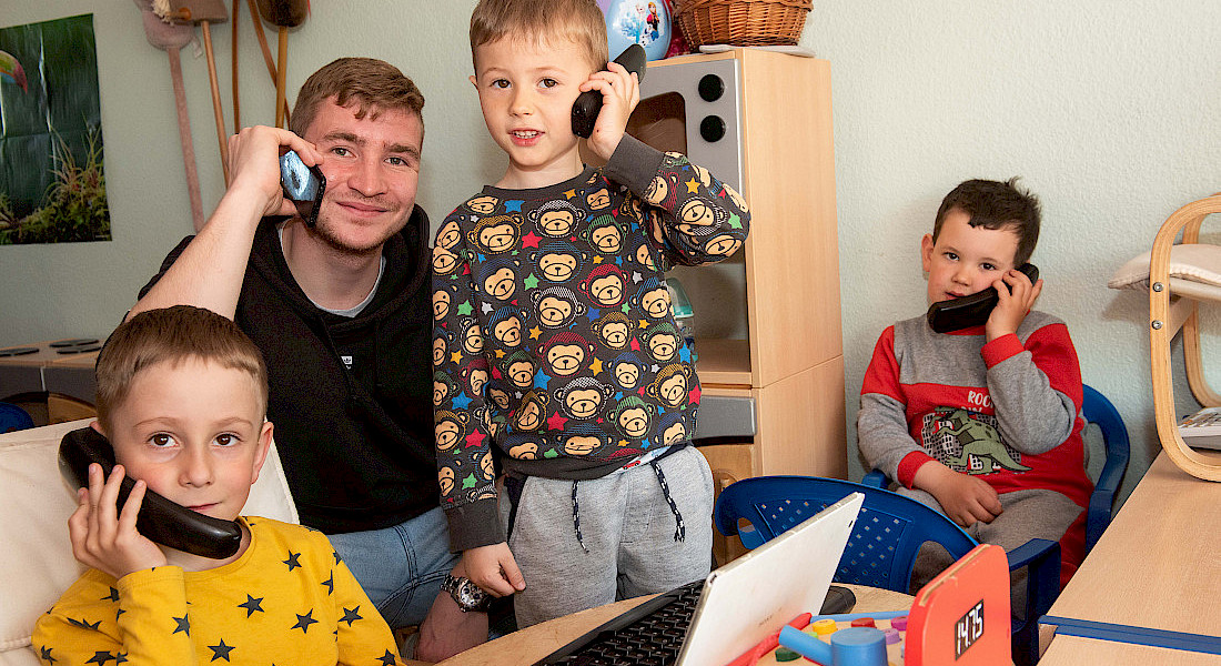 Kinder sitzen an einem Tisch und spielen mit Telefonhörern, ein Erwachsener ist dabei.