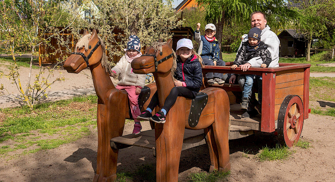 Kinder reiten auf Holzpferden und Kutsche, eine erwachsene Person begleitet sie.