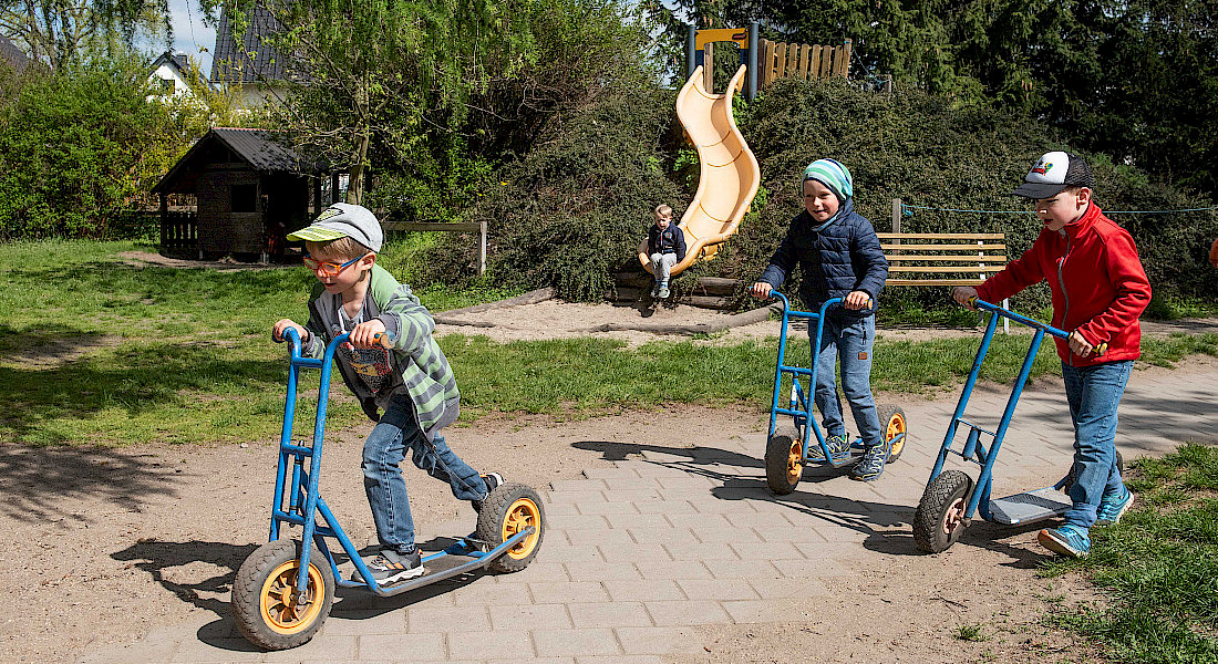 Kinder fahren draußen auf Rollern, weitere Kinder sind im Hintergrund zu sehen.