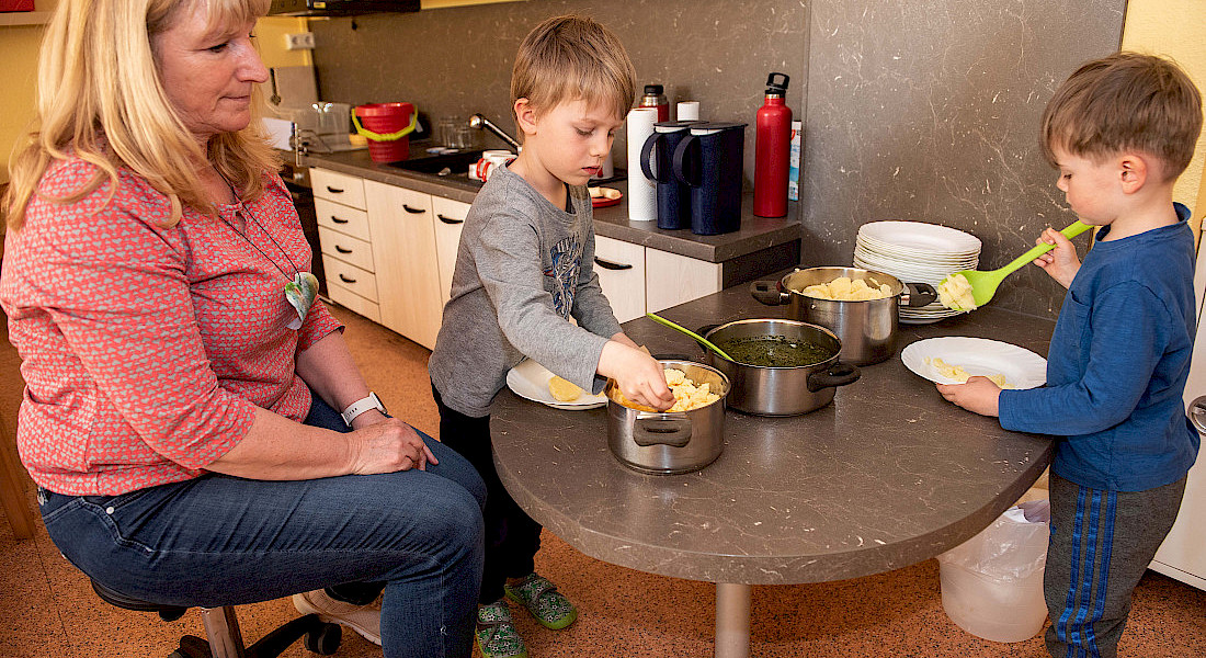 Zwei Kinder schöpfen Essen aus einem Topf, eine erwachsene Person begleitet sie.