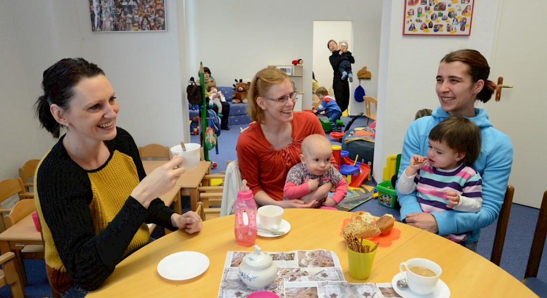 Eltern sitzen  mit ihren Babys an Kaffeetisch und lächeln.