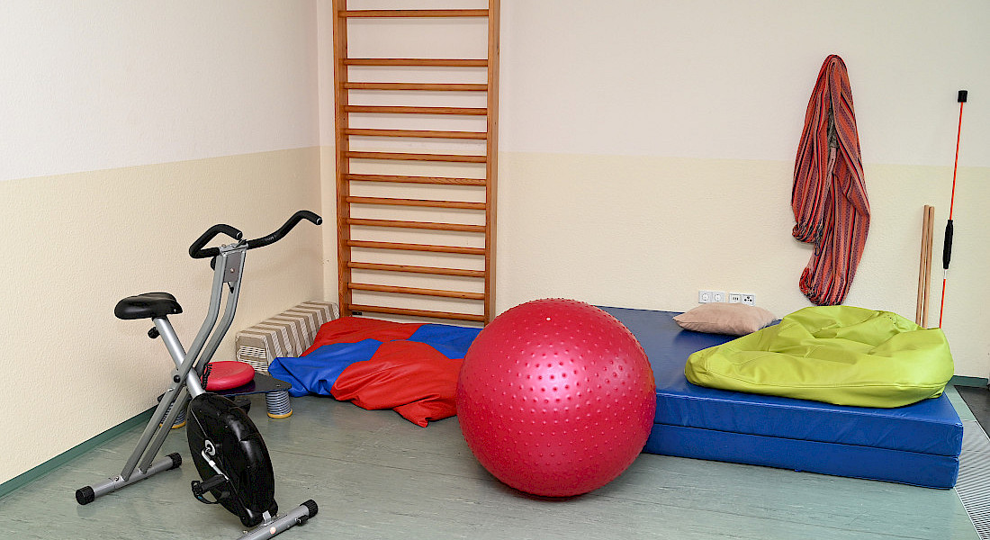 Blick in einen Bewegungsraum mit Heimtrainer, Ball, Sprossenwand und Matten.