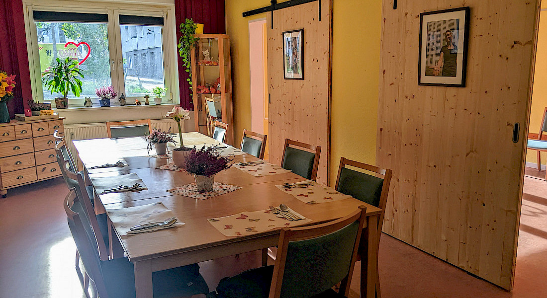 Blick in einen gemütlichen Raum zum Beisammensein. mit langem Holztisch und rollbaren Holztüren und freundlichen Bildern und Pflanzen.