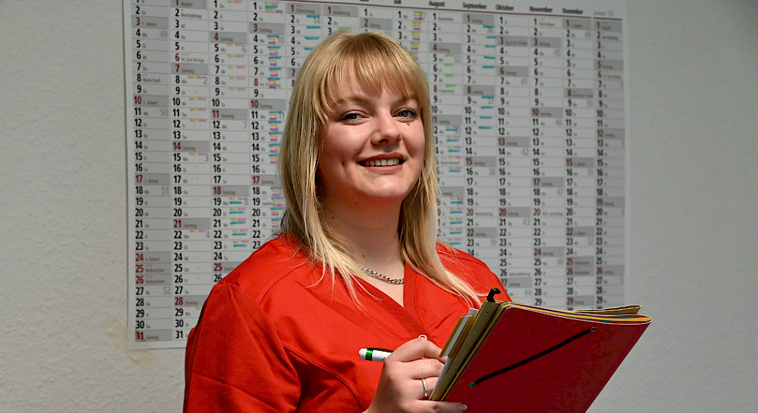 Person mit Mappe und Stift in der Hand lächelt in die Kamera, Kalender an der Wand.