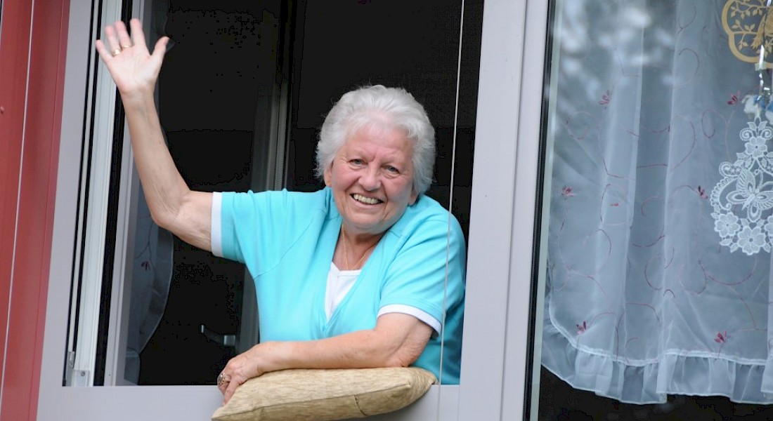 Eine Frau lächelt und winkt aus ihrem offenen Fenster, auf der Fensterbank hat sie ein Kissen.