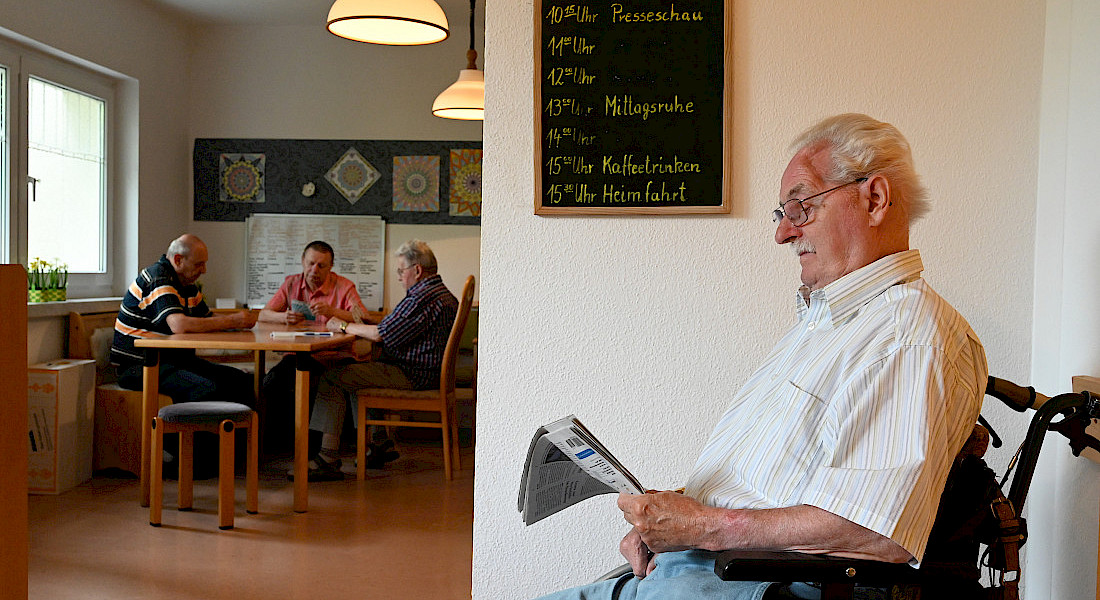 Im Vordergrund sitzt eine ältere Person im Rollstuhl und liest Zeitung, im Hintergrund sitzen drei kartenspielende ältere Personen an einem Tisch,