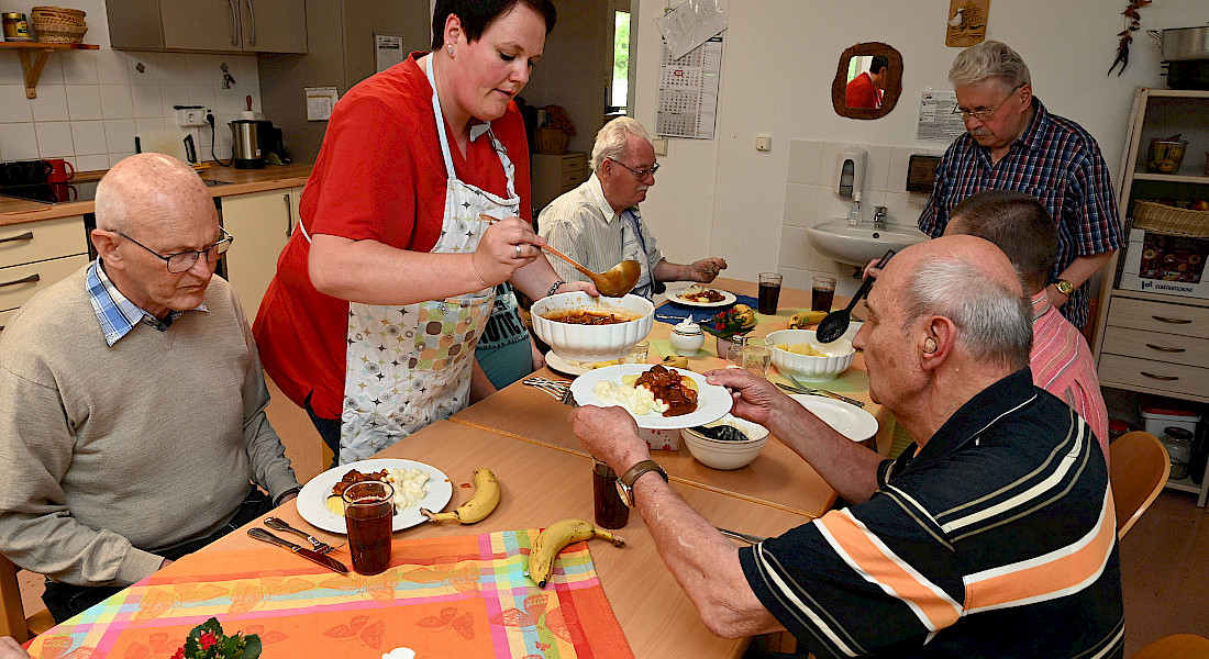 Eine Pflegekraft serviert ältereb Person das Mittagessen am Küchentisch in der Küche.