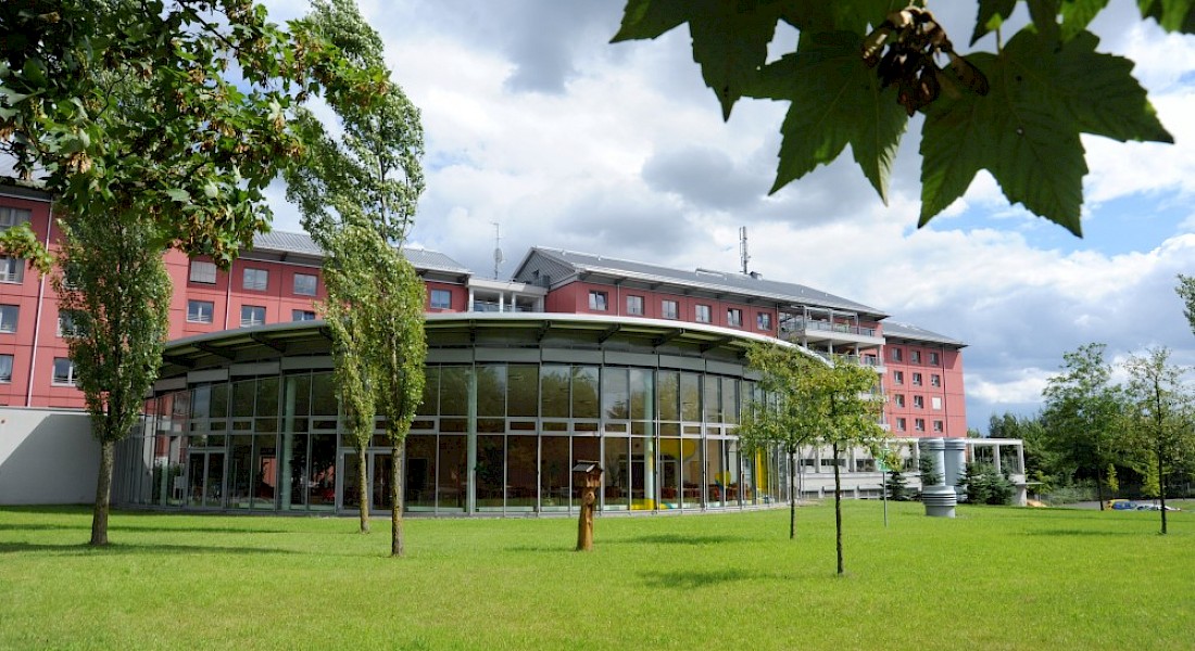 Blick auf rotes Gebäude mit großem verglastem Saal, davor eine große Grünfläche.
