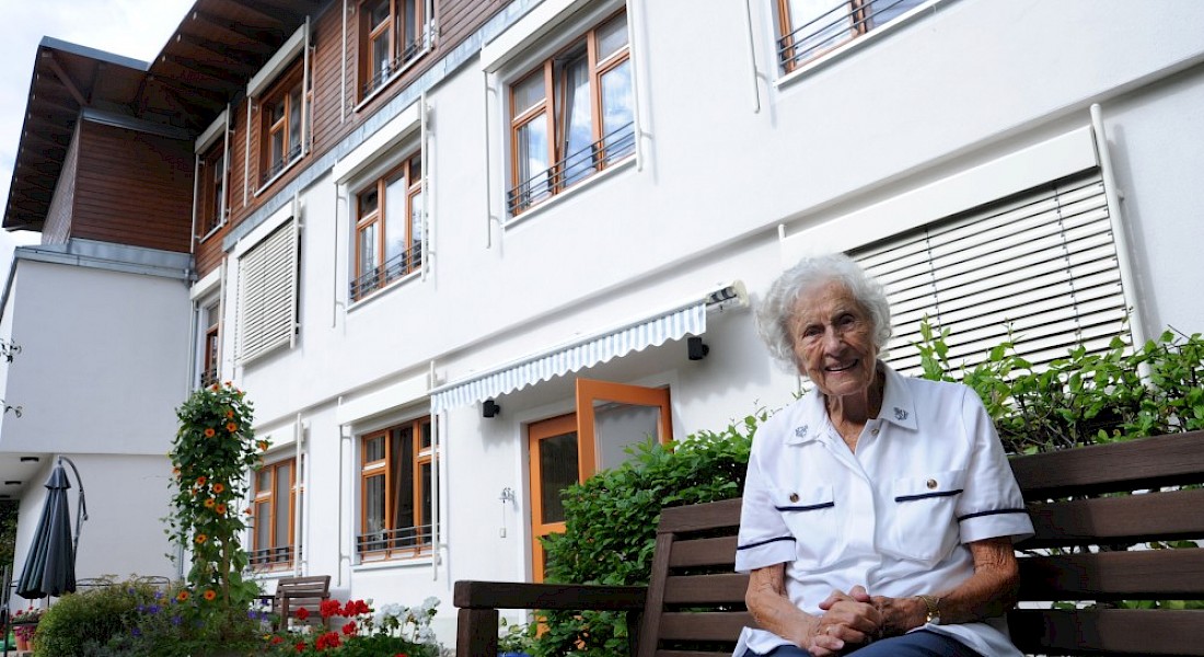 Eine Frau sitzt vor einem weißen Haus auf einer Bank.