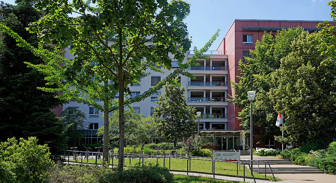 Außenansicht der mehrstöckigen Einrichtung mit Bäumen und Park im Vordergrund.