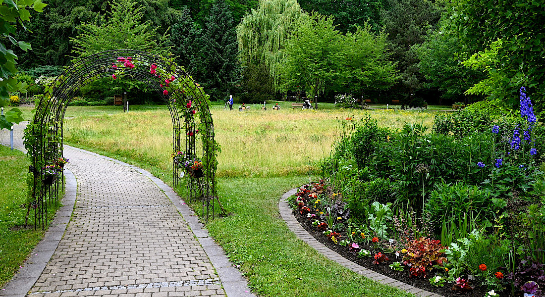 Gepflegter Gartenweg mit Rosenbogen und Blumenbeet auf einer Wiese.