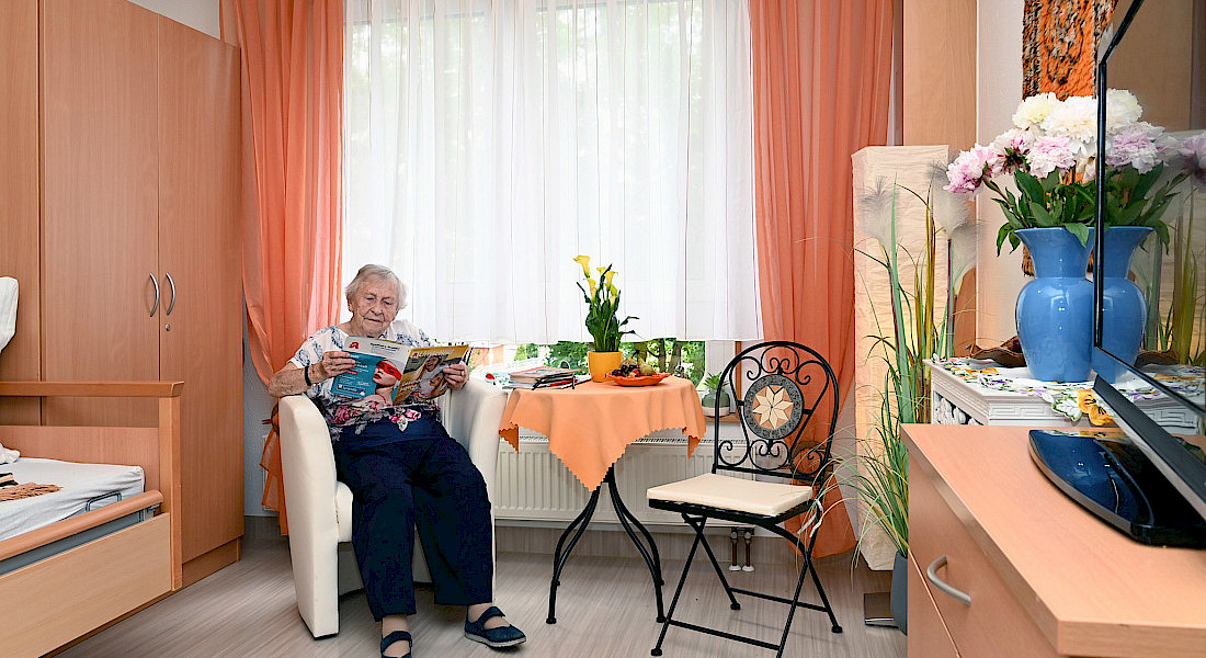 Eine ältere Person liest im Sessel neben einem kleinen runden Tisch in einem hellen und gemütlich eingerichtetem Pflegezimmer.