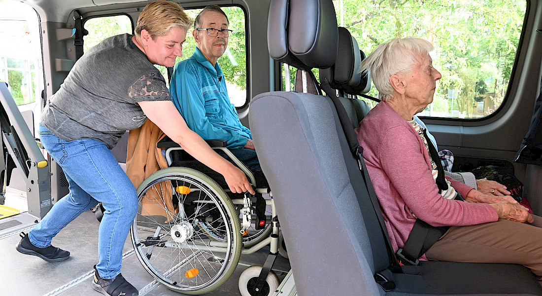 Eine Person hilft beim Einsteigen einer Person im Rollstuhl in ein Transportfahrzeug.