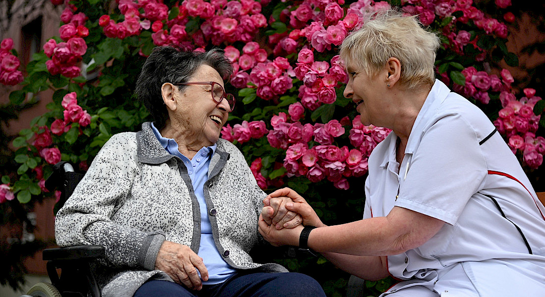 Eine ältere Person im Rollstuhl unterhält sich mit einer Pflegekraft im Garten vor blühenden Rosen und lachen einander an.