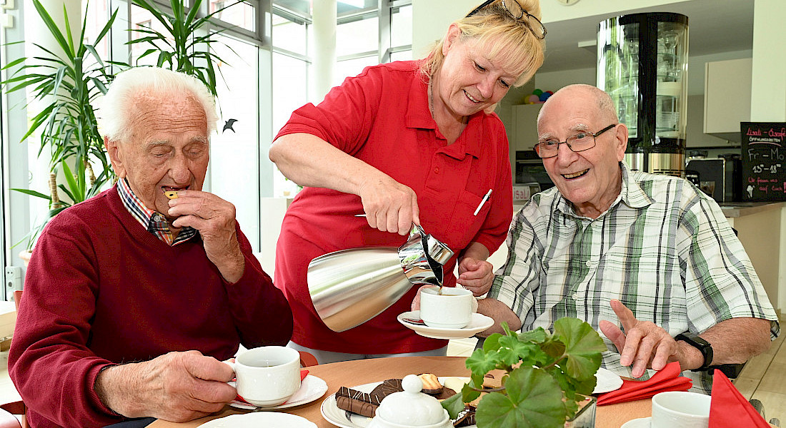 Zwei ältere Personen sitzen lächelnd bei Kaffee und Gebäck an einem Tisch, während die Pflegekraft Kaffee nachschenkt.