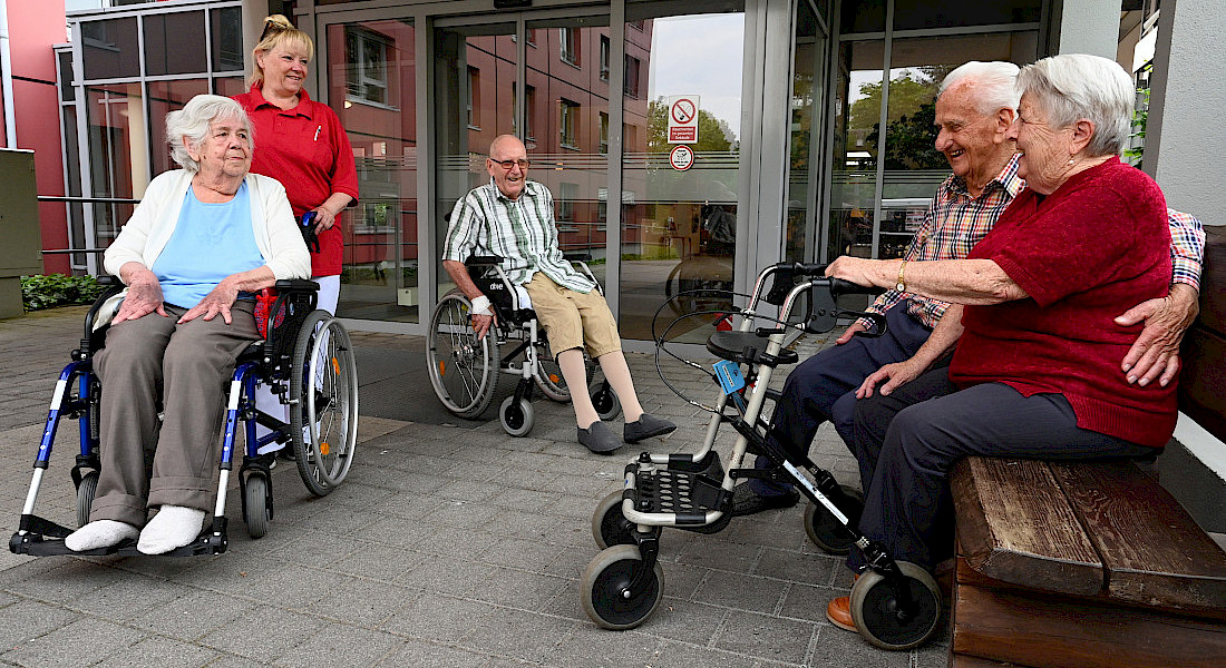 Gruppe älterer Personen, teils im Rollstuhl und auf einer Parkbank, vor dem Eingang eines Pflegeheims mit einer Pflegekraft.
