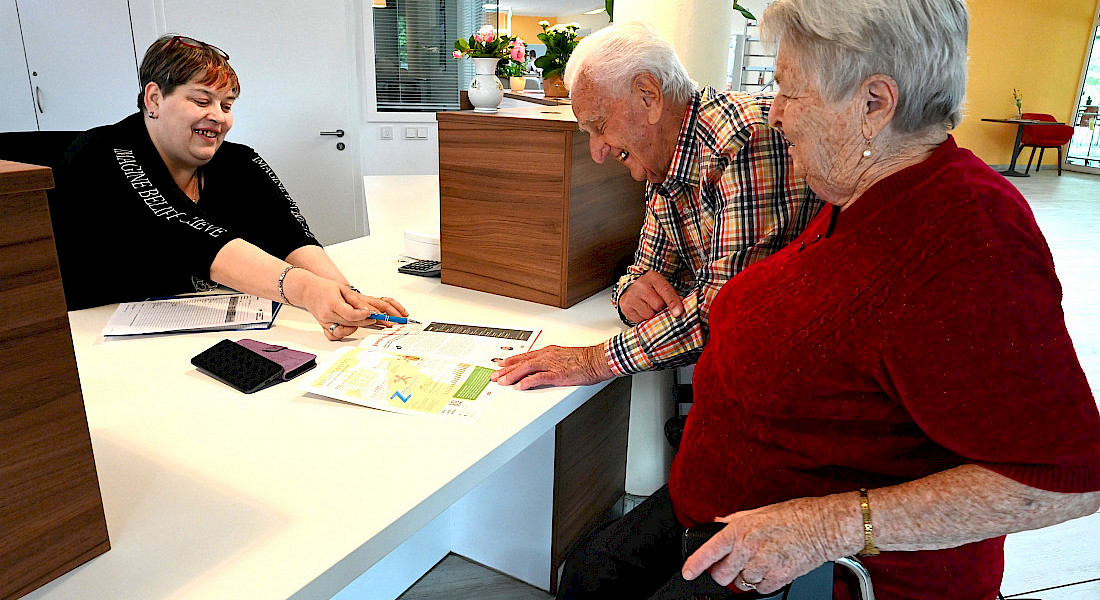 Rezeptionspersonal hilft am Empfangstresen zwei älteren Personen mit Informationsmaterial.