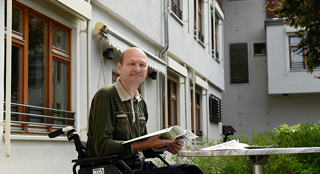 Eine Person im Rollstuhl sitzt mit Zeitung im Innenhof der Einrichtung.