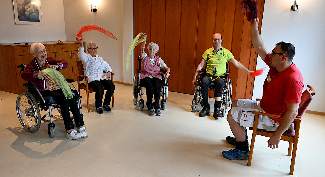 Gruppe von vier älteren Personen in Rollstühlen, macht Gymnastik mit einer Pflegekraft. Sie üben mit roten Tüchern.