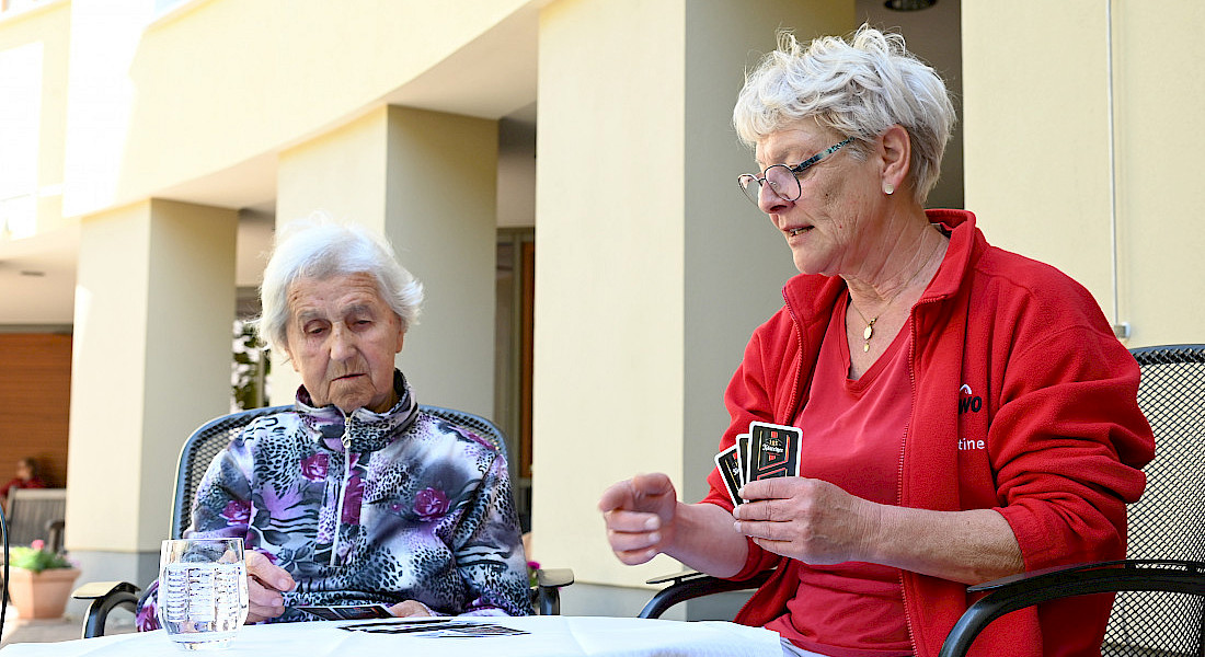 Eine Pflegekraft spielt mit einer älteren Person Karten an einem Tisch im Freien.