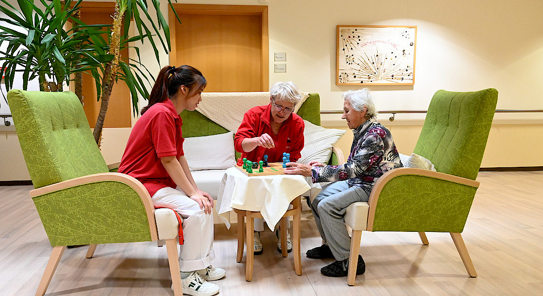 Zwei Pflegekräfte spielen mit einer älteren Person Mensch-Ärgere-Dich-Nicht an einem Tisch mit gemütlichen Sesseln in einem freundlich gestalteten Flur.