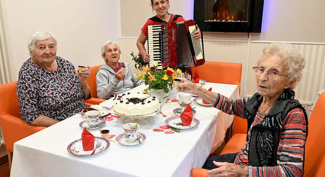 Drei ältere Personen feiern an einem gedeckten Tisch mit Kuchen, während eine Person Akkordeon spielt.