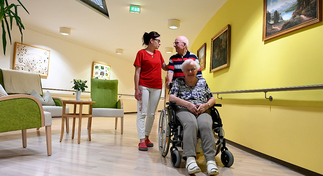 Zwei Pflegekräfte begleiten ein ältere Person im Rollstuhl durch einen hellen Gang.